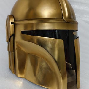May include: A gold-colored helmet with a visor. The helmet has a rounded top and angular features, with a dark visor area. The helmet is made of a shiny, metallic material. The design is reminiscent of a Mandalorian helmet.