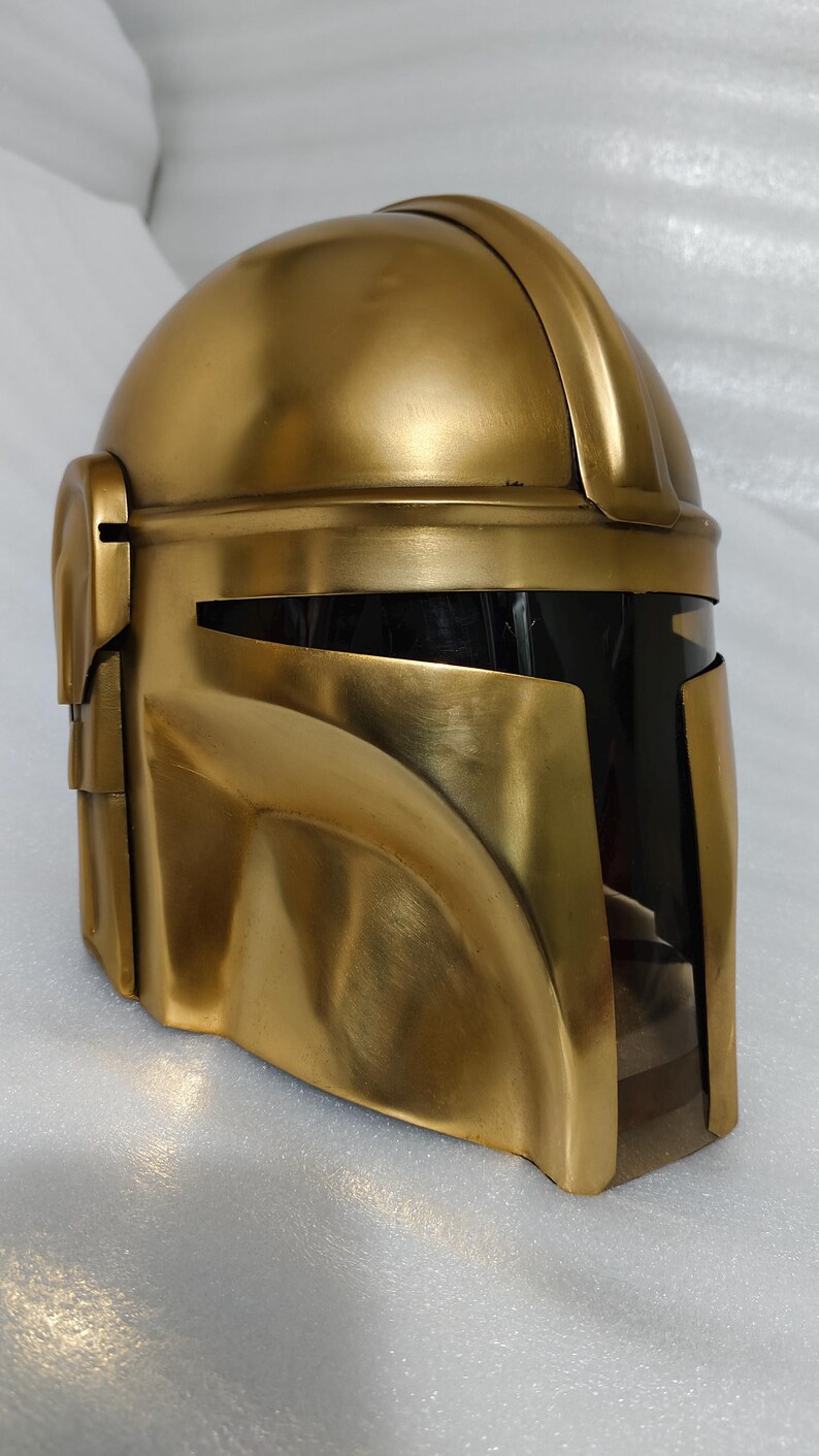 May include: A gold-colored helmet with a visor. The helmet has a rounded top and angular features, with a dark visor area. The helmet is made of a shiny, metallic material. The design is reminiscent of a Mandalorian helmet.