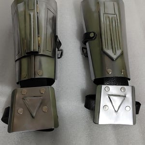 Puede incluir: Dos protectores de brazo plateados con correas de cuero negro. Cada protector tiene un dise&ntilde;o rectangular con formas geom&eacute;tricas y remaches. Los protectores son probablemente para disfraces o cosplay.