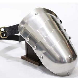 May include: A polished steel pauldron, a piece of medieval armor, with a segmented design and riveted construction. It features a black leather strap with a brass buckle for attachment. The pauldron is displayed on a wooden stand.