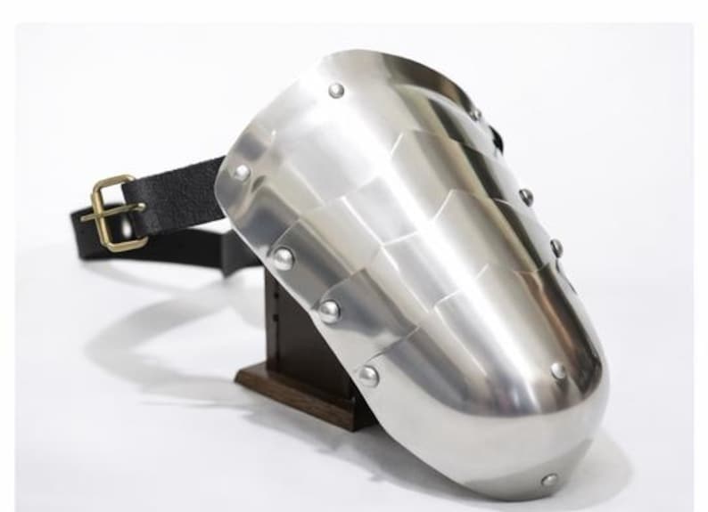 May include: A polished steel pauldron, a piece of medieval armor, with a segmented design and riveted construction. It features a black leather strap with a brass buckle for attachment. The pauldron is displayed on a wooden stand.