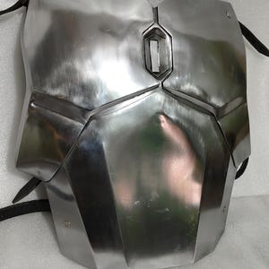 Puede incluir: Una placa de pecho plateada con un dise&ntilde;o geom&eacute;trico. La placa tiene una abertura rectangular central y est&aacute; asegurada con correas negras. La superficie met&aacute;lica refleja la luz, resaltando la construcci&oacute;n angular de la armadura.