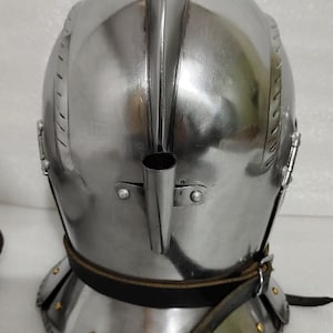 May include: A polished steel helmet with a prominent central ridge and a tubular attachment. The helmet features a neck guard and a leather strap with rivets. This medieval-style helmet is designed for protection.