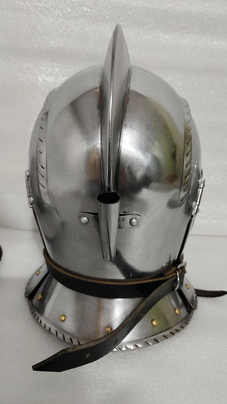 May include: A polished steel helmet with a prominent central ridge and a tubular attachment. The helmet features a neck guard and a leather strap with rivets. This medieval-style helmet is designed for protection.