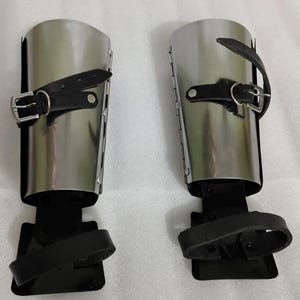 Puede incluir: Par de grebas de metal pulido con correas y hebillas de cuero negro. La armadura de la pierna tiene un dise&ntilde;o cil&iacute;ndrico y est&aacute; dise&ntilde;ada para proteger la parte inferior de la pierna. Las grebas se muestran sobre una superficie blanca.