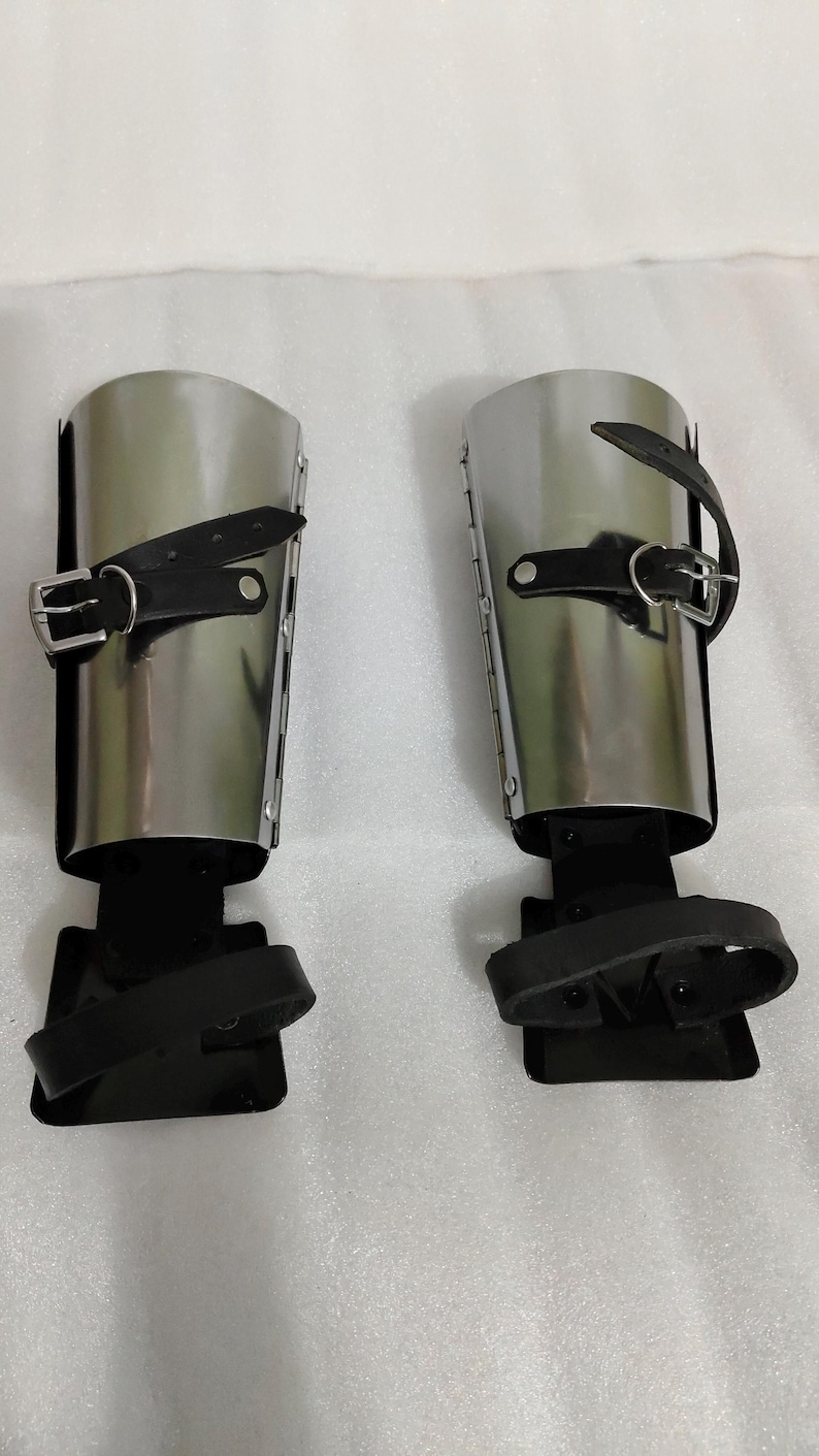 Puede incluir: Par de grebas de metal pulido con correas y hebillas de cuero negro. La armadura de la pierna tiene un dise&ntilde;o cil&iacute;ndrico y est&aacute; dise&ntilde;ada para proteger la parte inferior de la pierna. Las grebas se muestran sobre una superficie blanca.