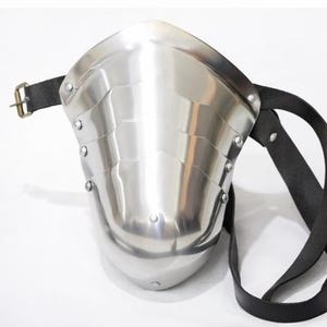 May include: A polished steel shoulder armor piece with a riveted design and a black leather strap. The armor has a curved shape and a buckle closure. The metal reflects light, creating a shiny appearance. This medieval-style armor is designed for protection.