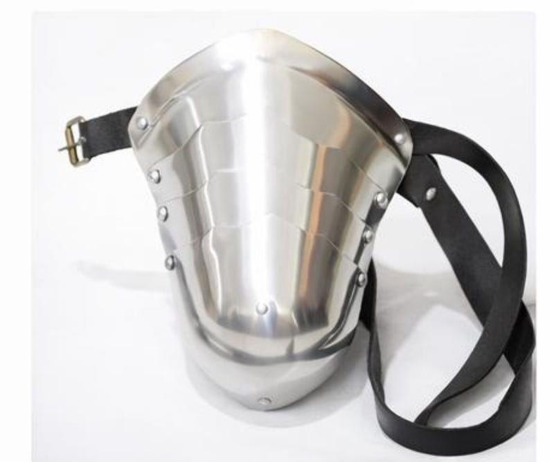May include: A polished steel shoulder armor piece with a riveted design and a black leather strap. The armor has a curved shape and a buckle closure. The metal reflects light, creating a shiny appearance. This medieval-style armor is designed for protection.
