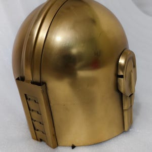 May include: A gold-colored helmet with a rounded top and a structured design. The helmet features a detailed side panel and a chin guard. The metallic finish gives it a futuristic or historical aesthetic.