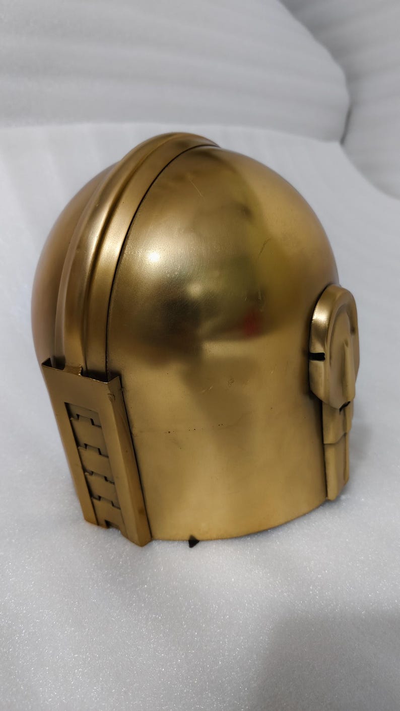 May include: A gold-colored helmet with a rounded top and a structured design. The helmet features a detailed side panel and a chin guard. The metallic finish gives it a futuristic or historical aesthetic.