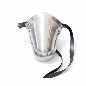 May include: A polished metal arm guard with a riveted design and a black leather strap. The armor piece is designed to protect the forearm and is secured with a buckle. The item is set against a white background.