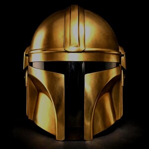 May include: A gold-colored Mandalorian helmet with a sleek, metallic finish. The helmet features a T-shaped visor and angular design, set against a black background. The helmet's surface reflects light, highlighting its detailed craftsmanship.