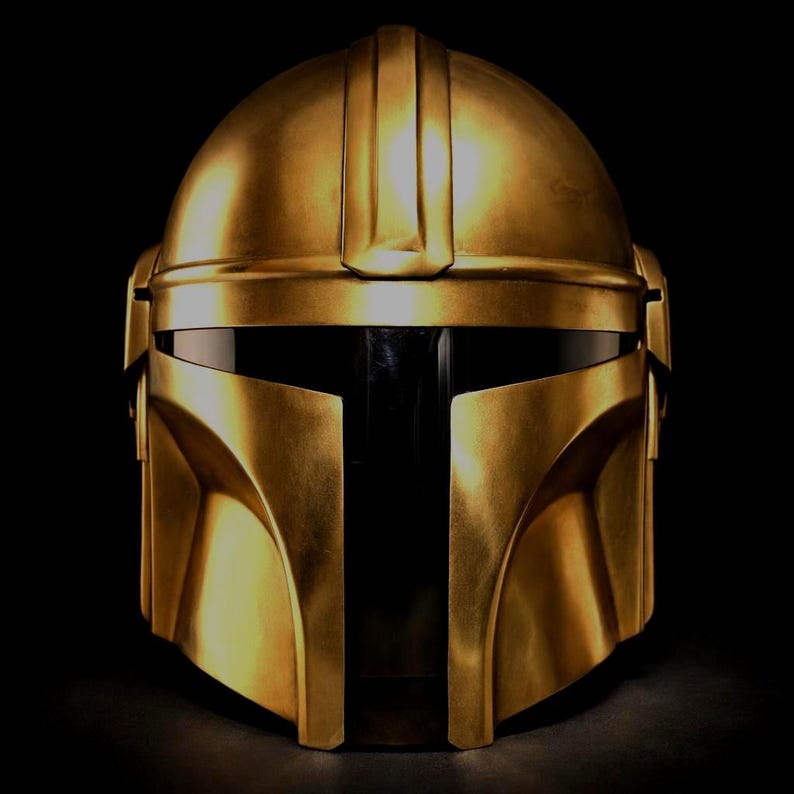May include: A gold-colored Mandalorian helmet with a sleek, metallic finish. The helmet features a T-shaped visor and angular design, set against a black background. The helmet's surface reflects light, highlighting its detailed craftsmanship.