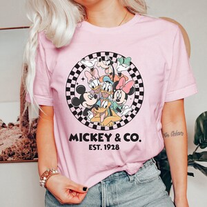 Puede incluir: Camiseta rosa claro con un dise&ntilde;o de Mickey Mouse, Minnie Mouse, Donald Duck, Daisy Duck, Goofy y Pluto. El dise&ntilde;o est&aacute; dentro de un c&iacute;rculo a cuadros blanco y negro con el texto "Mickey & Co. Est. 1928".