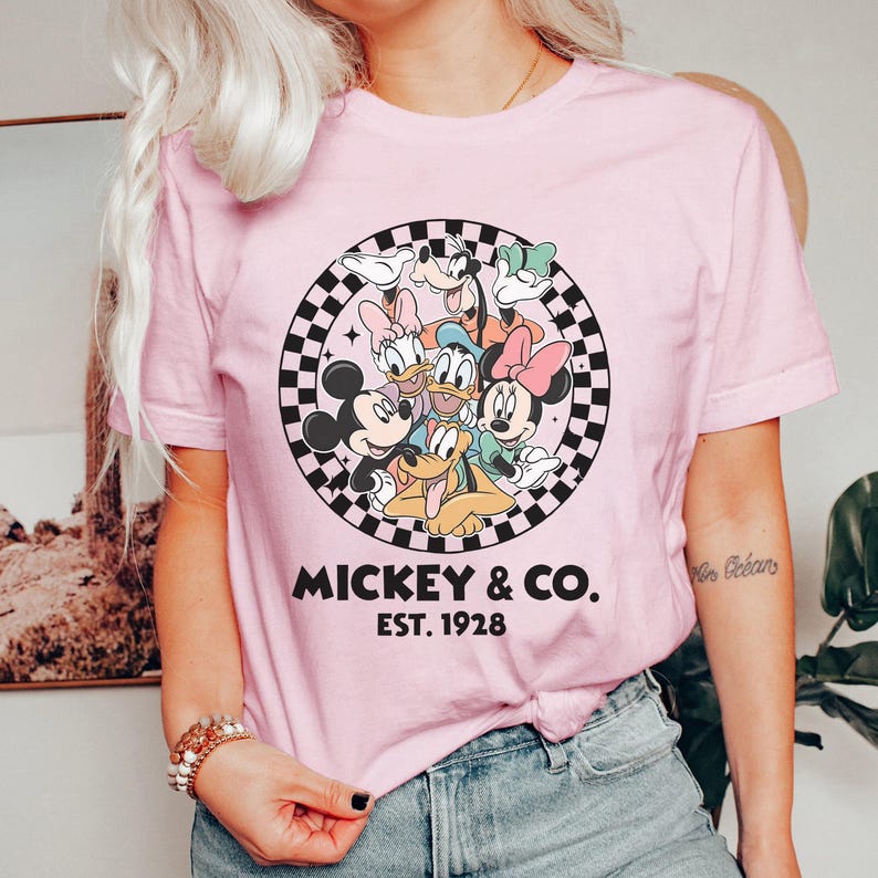 Puede incluir: Camiseta rosa claro con un dise&ntilde;o de Mickey Mouse, Minnie Mouse, Donald Duck, Daisy Duck, Goofy y Pluto. El dise&ntilde;o est&aacute; dentro de un c&iacute;rculo a cuadros blanco y negro con el texto "Mickey & Co. Est. 1928".