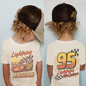 May include: A child wearing a cream-coloured t-shirt and a brown trucker hat. The front of the shirt features a graphic of Lightning McQueen from the film Cars. The back of the shirt displays the number 95 with flames and the words "Lightning McQueen".