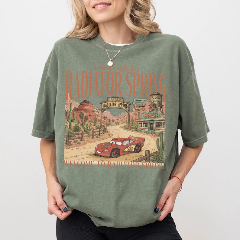 May include: Olive green t-shirt featuring a graphic of Radiator Springs from the movie Cars. The design includes a red race car, desert scenery, and the text "Radiator Spring" and "Welcome to Radiator Spring".
