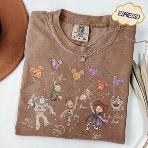 Puede incluir: Camiseta color espresso con personajes de Toy Story de Disney sosteniendo globos con forma de Mickey Mouse. La camiseta incluye a Buzz Lightyear, Woody, Jessie y Mr. Potato Head. La camiseta tiene la etiqueta de la marca Comfort Colors y un collar plateado.