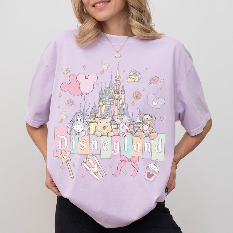 Puede incluir: Camiseta lila claro con un gr&aacute;fico de Disneyland. El dise&ntilde;o incluye un castillo, globos de Mickey Mouse y personajes de Disney. La palabra "Disneyland" est&aacute; impresa en colores pastel.