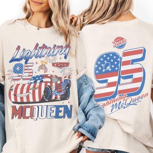 Może przedstawiać: Kremowy t-shirt z grafiką Lightning McQueen w motywy flagi amerykańskiej, numerem 95 i napisem Lightning McQueen. Z tyłu znajduje się duża liczba 95 z pasami flagi oraz logo Rust-eze w kolorze czerwonym i niebieskim.