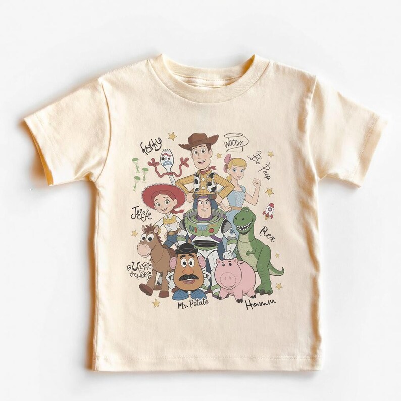 May include: A cream-colored short-sleeve t-shirt featuring a graphic print of Toy Story characters including Woody, Buzz Lightyear, Jessie, Bullseye, Rex, Hamm, Mr. Potato Head, Forky, and Bo Peep, with their names written in a cursive script.