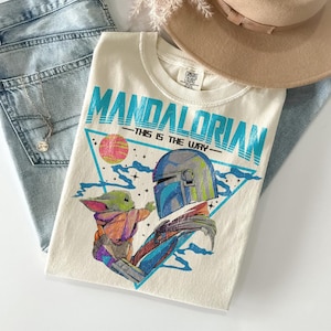 May include: Cream t-shirt with a graphic of The Mandalorian and Grogu, and the text "MANDALORIAN - THIS IS THE WAY" in teal. The design includes a helmet and a baby character. The shirt is on blue jeans and a tan hat. The shirt is made of cotton.