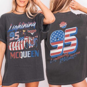 Może przedstawiać: Ciemnoszary t-shirt z kr&oacute;tkim rękawem z grafiką Lightning McQueen w amerykański wz&oacute;r flagi. Z przodu znajduje się napis Lightning McQueen i 95. Z tyłu widnieje duża liczba 95 z pasami flagi oraz logo Rust-eze.