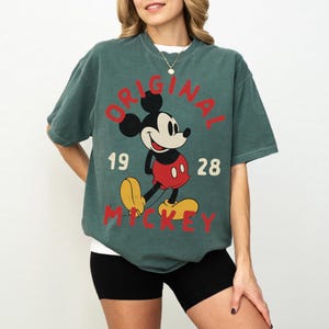 May include: Green t-shirt featuring a classic Mickey Mouse graphic. The design includes the words "ORIGINAL MICKEY" and the year "1928" in red. The shirt has short sleeves and a crew neck.