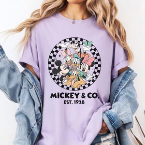Puede incluir: Camiseta lila claro con un gr&aacute;fico de Mickey Mouse y sus amigos dentro de un c&iacute;rculo a cuadros blanco y negro. El texto "MICKEY & CO. EST. 1928" est&aacute; impreso debajo.