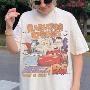 May include: Cream-colored t-shirt featuring a Halloween-themed graphic of characters from the movie Cars. The design includes the text "Radiator Springs" and "Trunk or Treat", with the number "95" in orange.