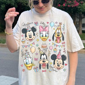 Puede incluir: Camiseta de color crema con los personajes de dibujos animados Mickey Mouse, Daisy Duck, Pluto, Donald Duck, Goofy y Minnie Mouse. El dise&ntilde;o incluye gorros de fiesta, globos, magdalenas y el texto "Mickey Mouse" y "Daisy Duck".