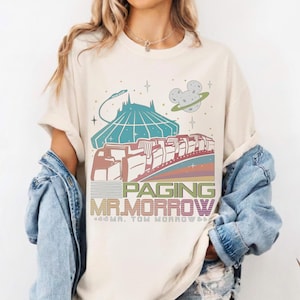 May include: Cream-coloured t-shirt with a retro graphic featuring a space-themed design. The graphic includes a futuristic building, a train, a planet with a Mickey Mouse head, and the text "Paging Mr. Morrow".