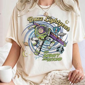 May include: A cream-colored t-shirt featuring a graphic of Buzz Lightyear in a space suit, surrounded by a blue spiral, planets, and stars. The text reads Buzz Lightyear and To Infinity and Beyond in a retro, curved, green and yellow font style.