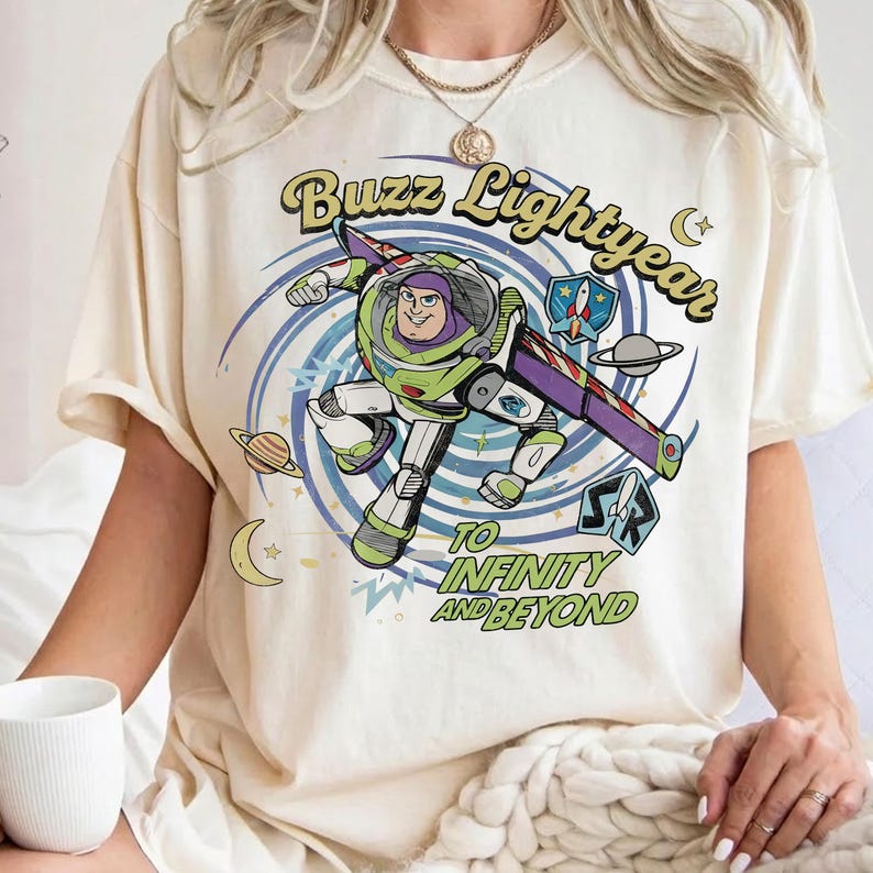 May include: A cream-colored t-shirt featuring a graphic of Buzz Lightyear in a space suit, surrounded by a blue spiral, planets, and stars. The text reads Buzz Lightyear and To Infinity and Beyond in a retro, curved, green and yellow font style.