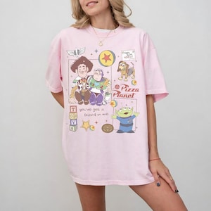 Pu&ograve; includere: T-shirt rosa chiaro con una grafica colorata dei personaggi di Toy Story. Il design include Woody, Buzz Lightyear, un alieno verde e il logo Pizza Planet. Il testo sulla maglietta recita "You've got a friend in me."