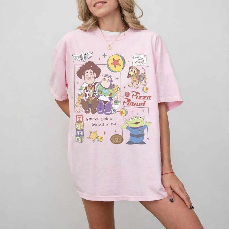 Pu&ograve; includere: T-shirt rosa chiaro con una grafica colorata dei personaggi di Toy Story. Il design include Woody, Buzz Lightyear, un alieno verde e il logo Pizza Planet. Il testo sulla maglietta recita "You've got a friend in me."