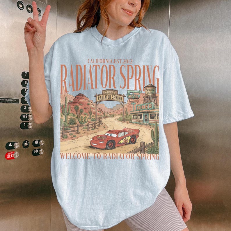 May include: Light blue t-shirt featuring a graphic of a red race car and the words "RADIATOR SPRING" and "WELCOME TO RADIATOR SPRING". The design includes a desert landscape with cacti and a diner. The text "CALIFORNIA.EST.2012" is also present.