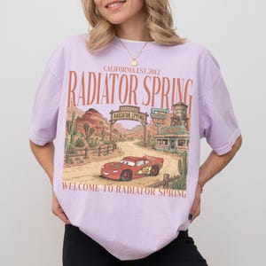 May include: A light purple t-shirt featuring a graphic of Radiator Springs from the movie Cars. The design includes a red race car, desert scenery, and the text "Radiator Spring" and "Welcome to Radiator Spring".