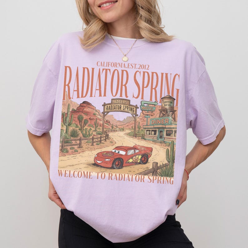 May include: A light purple t-shirt featuring a graphic of Radiator Springs from the movie Cars. The design includes a red race car, desert scenery, and the text "Radiator Spring" and "Welcome to Radiator Spring".