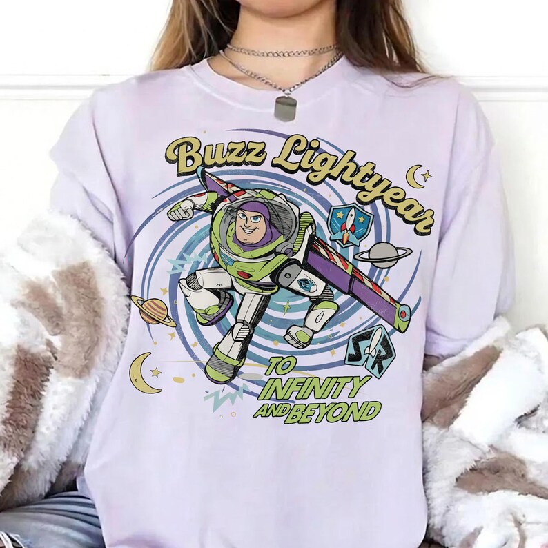 May include: A light purple t-shirt featuring a graphic of Buzz Lightyear in a space suit, surrounded by planets, stars, and a crescent moon. The text reads Buzz Lightyear and TO INFINITY AND BEYOND in a retro font style with a blue spiral background.