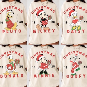 May include: A collection of six cream-colored t-shirts, each featuring a different Disney character wearing a red Santa hat and holiday sweater. The designs include Pluto, Mickey, Daisy, Donald, Minnie, and Goofy, with the text CHRISTMAS, the character name, and a year printed in red.