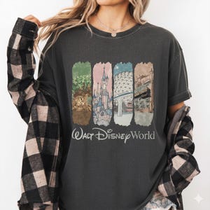 May include: Dark gray t-shirt featuring a colorful graphic of Walt Disney World landmarks. The design includes images of the Tree of Life, Cinderella Castle, Spaceship Earth, and Tower of Terror. The words "Walt Disney World" are printed below the graphic.