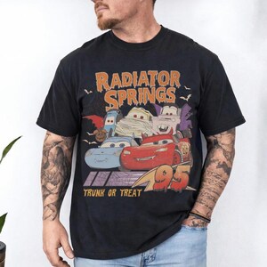 May include: Black t-shirt featuring a Halloween-themed graphic of characters from the movie Cars. The design includes the text "Radiator Springs" in orange, "Trunk or Treat", and the number "95".