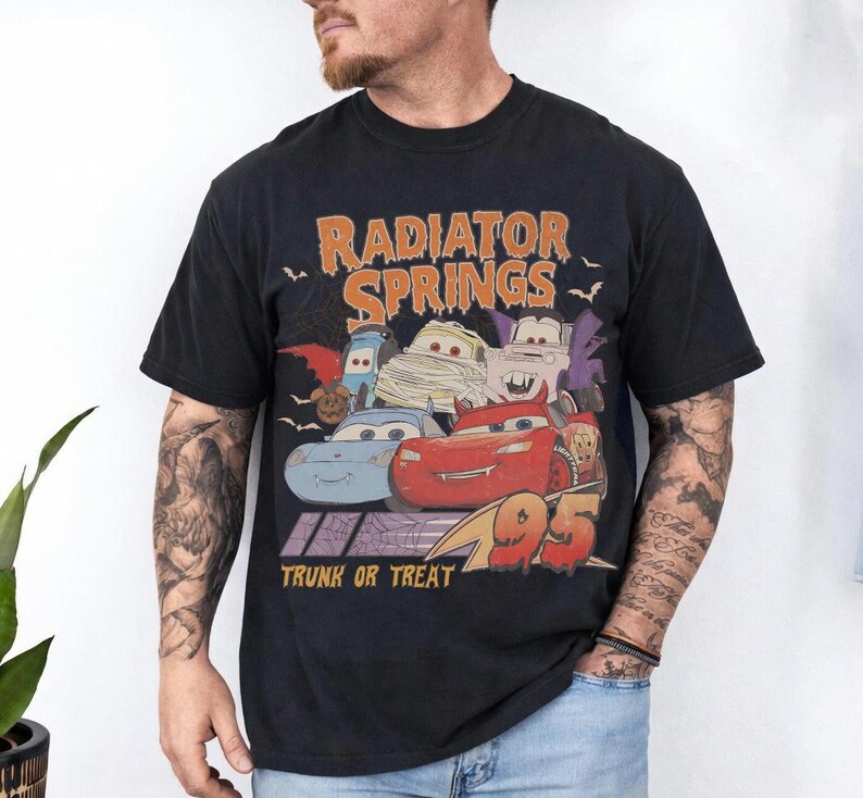 May include: Black t-shirt featuring a Halloween-themed graphic of characters from the movie Cars. The design includes the text "Radiator Springs" in orange, "Trunk or Treat", and the number "95".