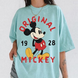 May include: A light blue t-shirt featuring a graphic of Mickey Mouse. The design includes the words "ORIGINAL MICKEY" in red, with the year "1928" below the word "ORIGINAL". Mickey Mouse is depicted in his classic red shorts and yellow shoes.