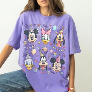 Puede incluir: Una camiseta lavanda con los personajes de dibujos animados Mickey Mouse, Daisy Duck, Pluto, Donald Duck, Goofy y Minnie Mouse. El dise&ntilde;o incluye gorros de fiesta, globos y gr&aacute;ficos de pastel de cumplea&ntilde;os. El texto "Mickey Mouse" y "Daisy Duck" tambi&eacute;n es visible.