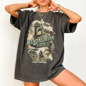 K&ouml;nnte beinhalten: Dunkelgraues T-Shirt mit einer Grafik aus der Star Wars-Serie "The Mandalorian and Grogu". Das Design enth&auml;lt Bilder des Mandalorianers, Grogu und den Text "THIS IS THE WAY".