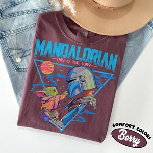 May include: Berry-coloured t-shirt featuring a graphic of The Mandalorian and Grogu, with the text "MANDALORIAN - THIS IS THE WAY" in blue. The shirt is part of the Comfort Colors collection.