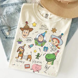 Peut inclure: T-shirt crème avec des personnages de dessins animés Woody, Buzz l'Éclair, Jessie, Slinky, Hamm et Rex. Le t-shirt porte l'inscription "Buzz l'Éclair" et les noms des personnages. Un chapeau beige et un collier argenté sont visibles.