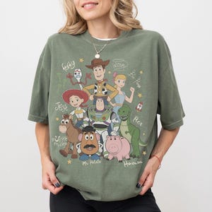 May include: An olive green short-sleeve t-shirt featuring a graphic print of Toy Story characters including Woody, Buzz Lightyear, Jessie, Bullseye, Mr. Potato Head, Hamm, Rex, Bo Peep, and Forky, with their names written in a cursive script font.